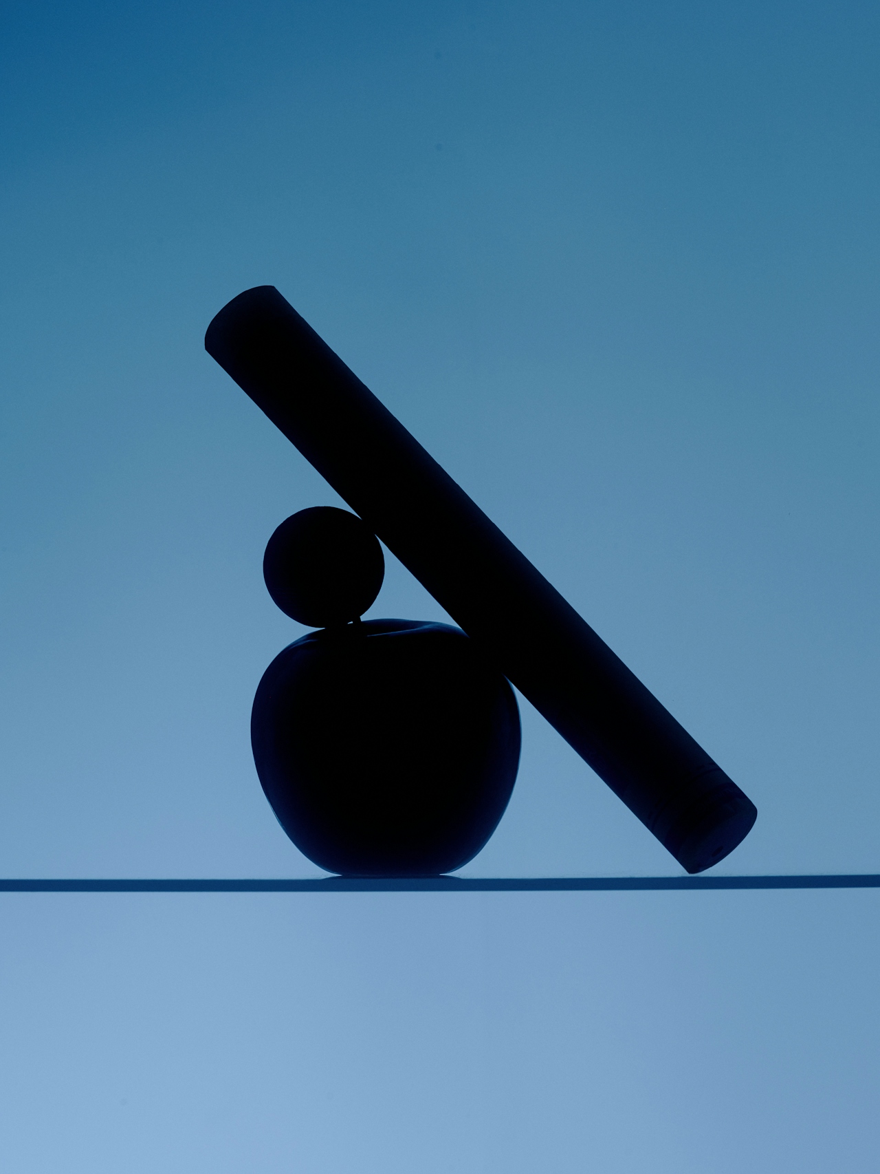 A dark cylinder leaning on an apple-shaped form, balanced over a horizon line at dusk.