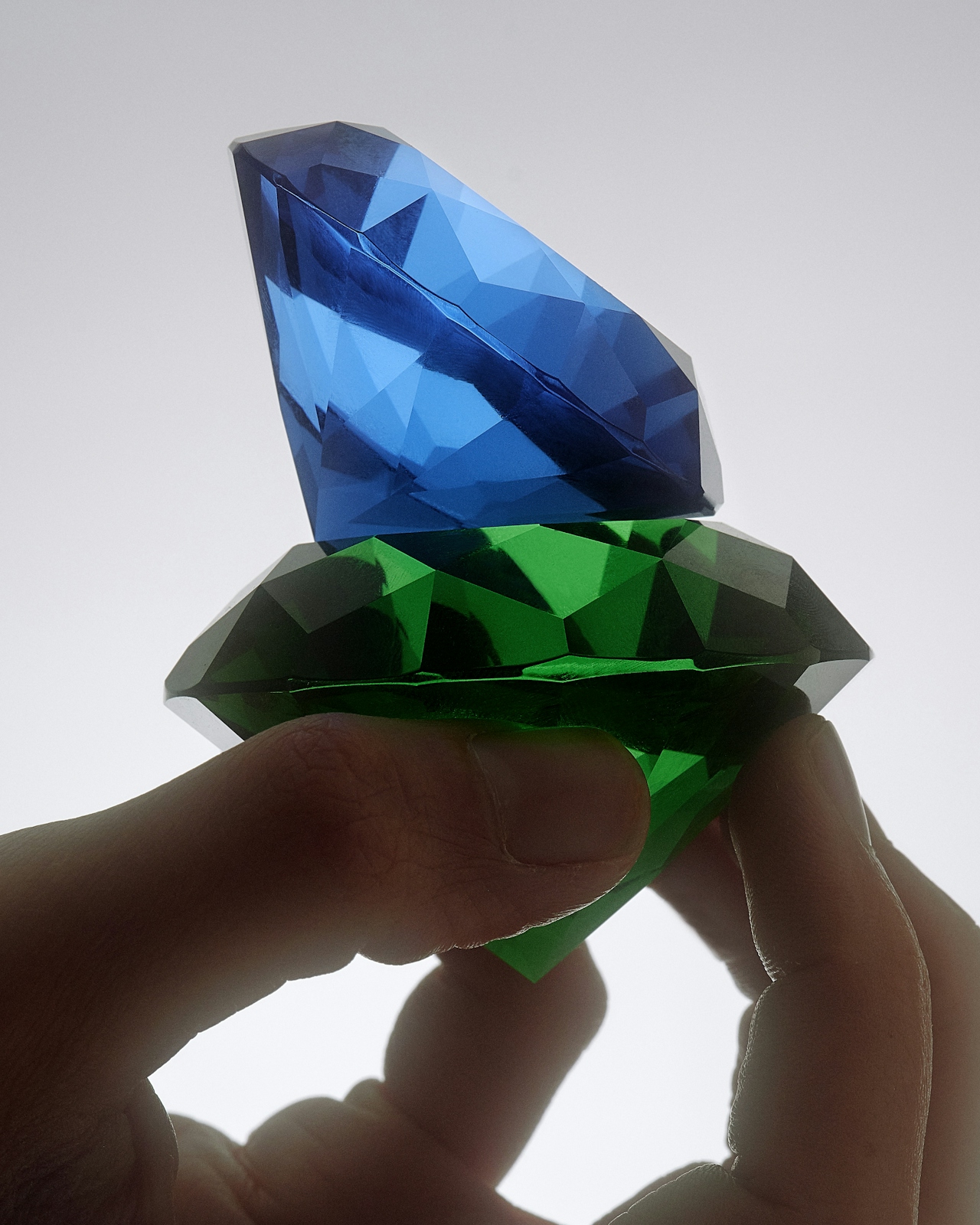 Fingertips holding a sapphire stacked atop an emerald against a soft white field.